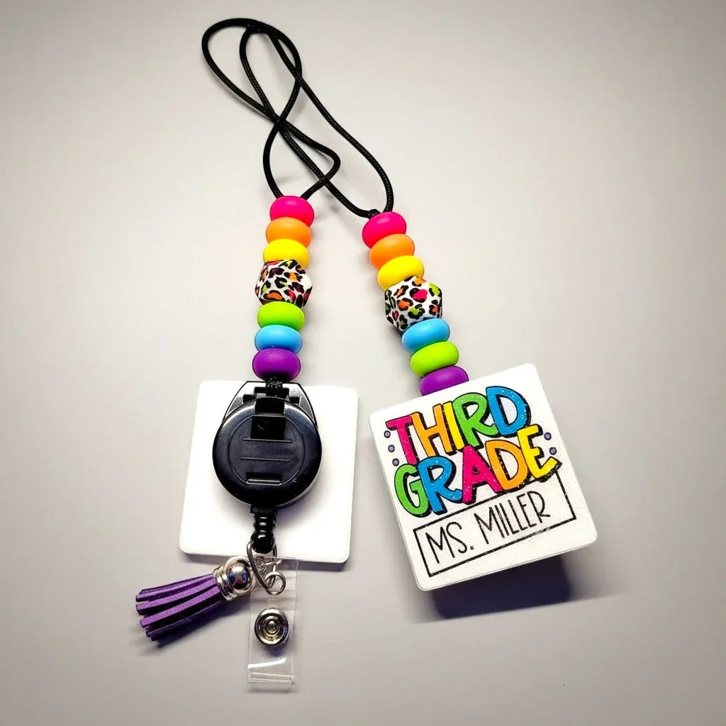 Mommie Peas Creations teacher lanyard with rainbow leopard print beads, a white badge holder, and a purple tassel.
