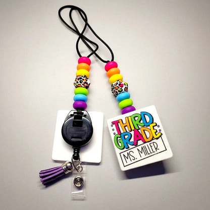 Mommie Peas Creations teacher lanyard with rainbow leopard print beads, a white badge holder, and a purple tassel.