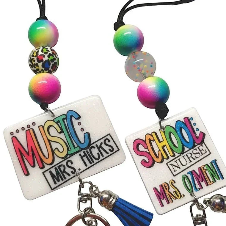 Mommie Peas Creations teacher lanyards with rainbow leopard print beads and custom name badges. One says "MUSIC MRS. HICKS" and the other "SCHOOL NURSE MRS. OZMINI".