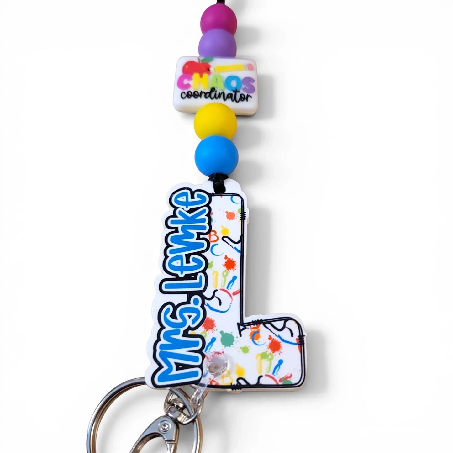 Mommie Peas Creations custom acrylic lanyard for Mrs. Levke, a special education teacher, featuring a white 'L' with colorful paint splatters and alphabet graphics, and a 'Chaos Coordinator' charm.