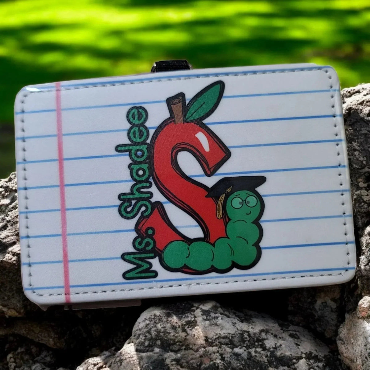 Mommie Peas Creations horizontal PU leather teacher badge holder with a red apple shaped letter S, a green caterpillar wearing a graduation cap, and the text "Ms. Shadee".