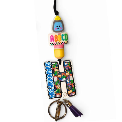Mommie Peas Creations personalized teacher lanyard with 'H' initial, ABC checkers pattern, pencil, smiley face beads, and purple tassel.