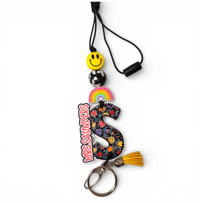 Mommie Peas Creations personalized teacher lanyard with 'S' initial, rainbow, smiley face, pencil beads, and yellow tassel.