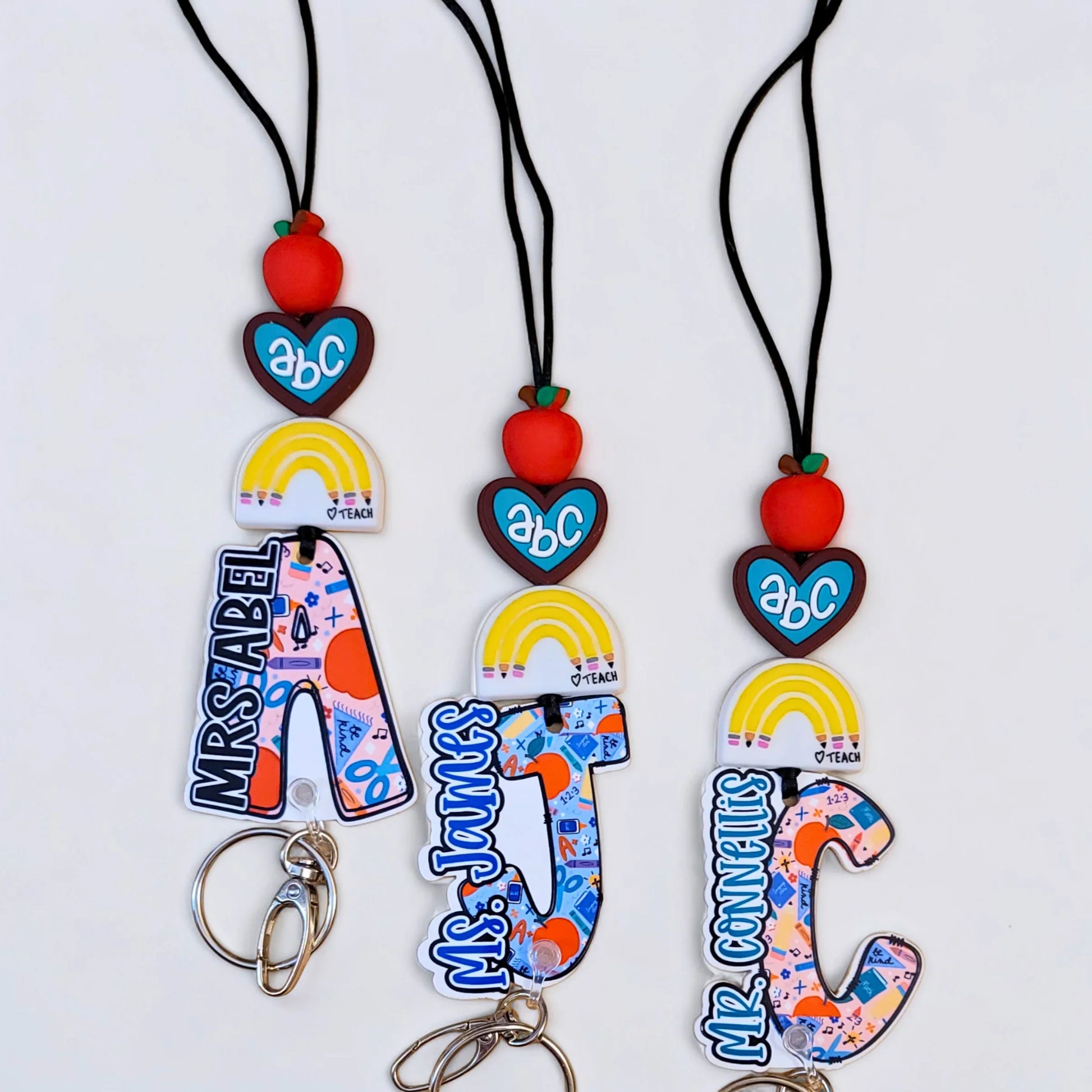 Mommie Peas Creations custom teacher lanyard with personalized initial, rainbow, apple, and ABC heart charms. Features a colorful school supply pattern.