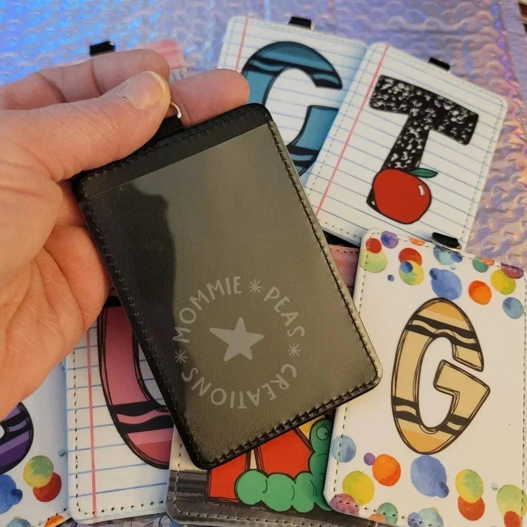 Mommie Peas Creations black faux leather vertical ID holder with clear window, surrounded by colorful badge holders with letters and school-themed designs.