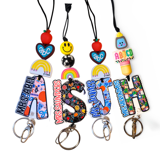 A collection of handmade teacher name badge holders with large acrylic initials. The lanyards feature various themes: a blue school-tool 'A', a black retro floral 'S', a striped 'J', and a colorful alphabet 'H', each with matching charms like apples, rainbows, and pencils.